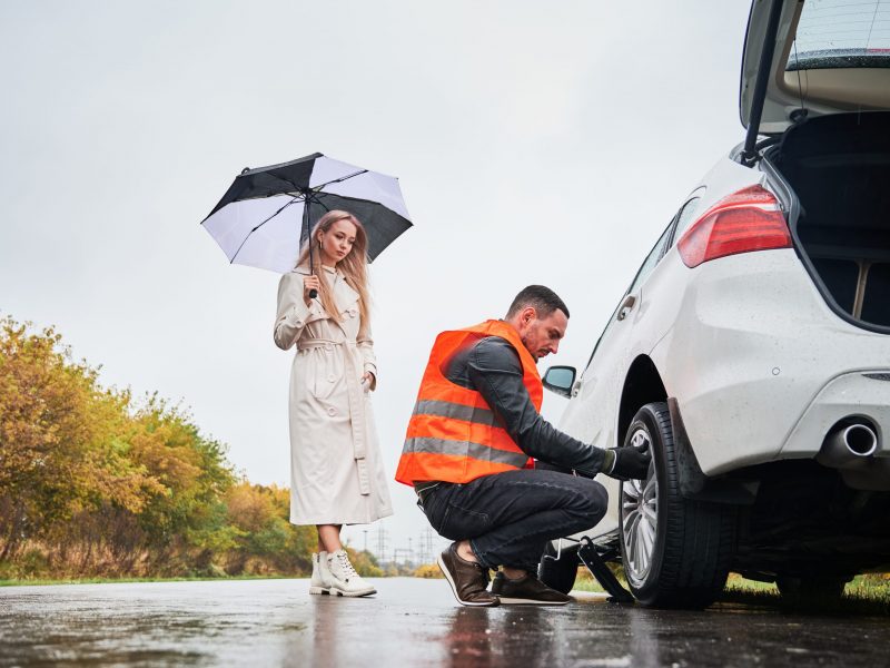 Roadside assistance worker replacing flat tire while beautiful woman in trench coat holding umbrella. Male auto mechanic changing flat tire on woman car the road. Concept of emergency road service.